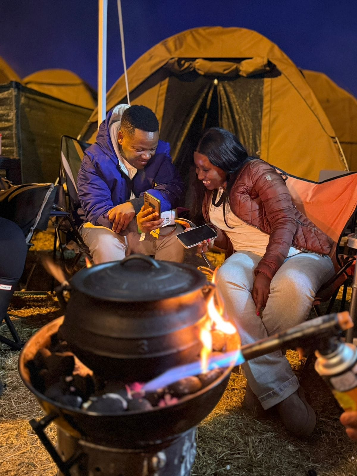 Two campers smiling together beside a cooking fire and canvas tents at night